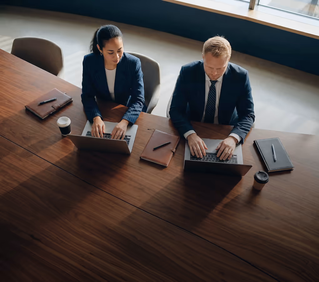 Two business professionals typing on laptops while sitting side by side at a conference table.