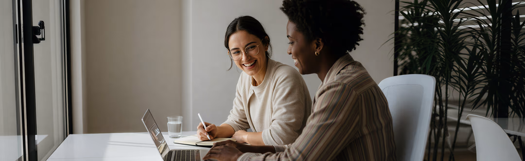 Two women smiling and collaborating at a laptop in a bright modern office with plants.