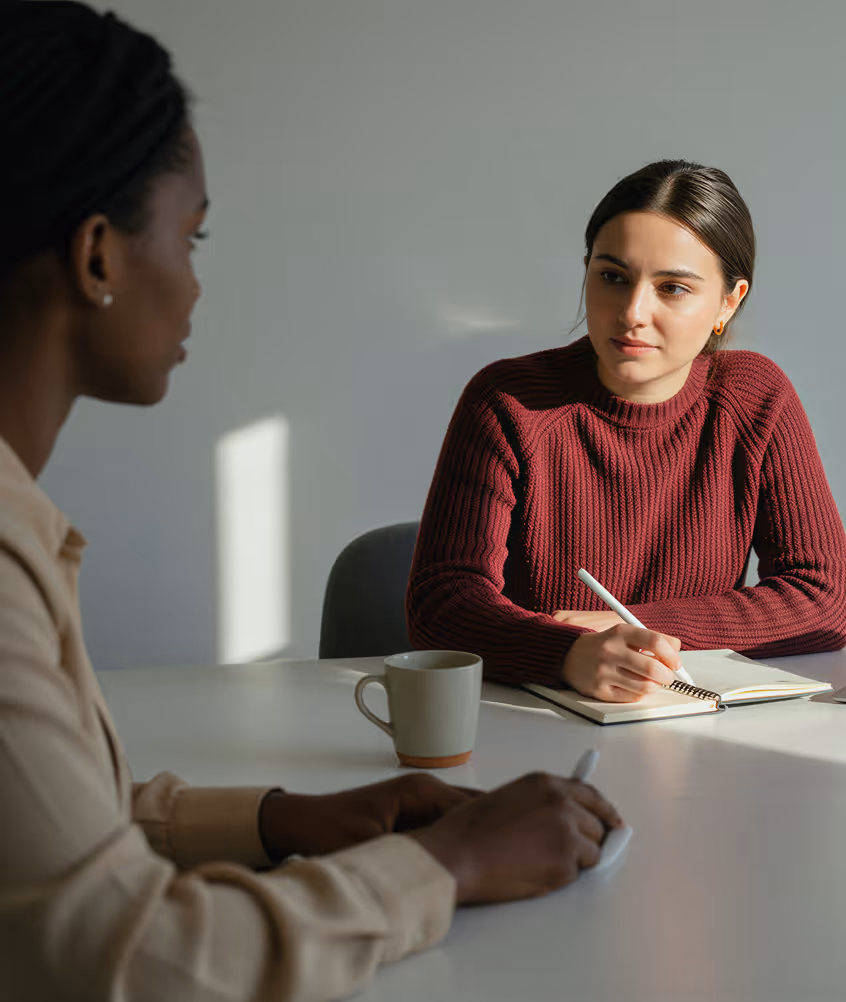 Two women sitting at a table having a serious conversation, one taking notes in a notebook.