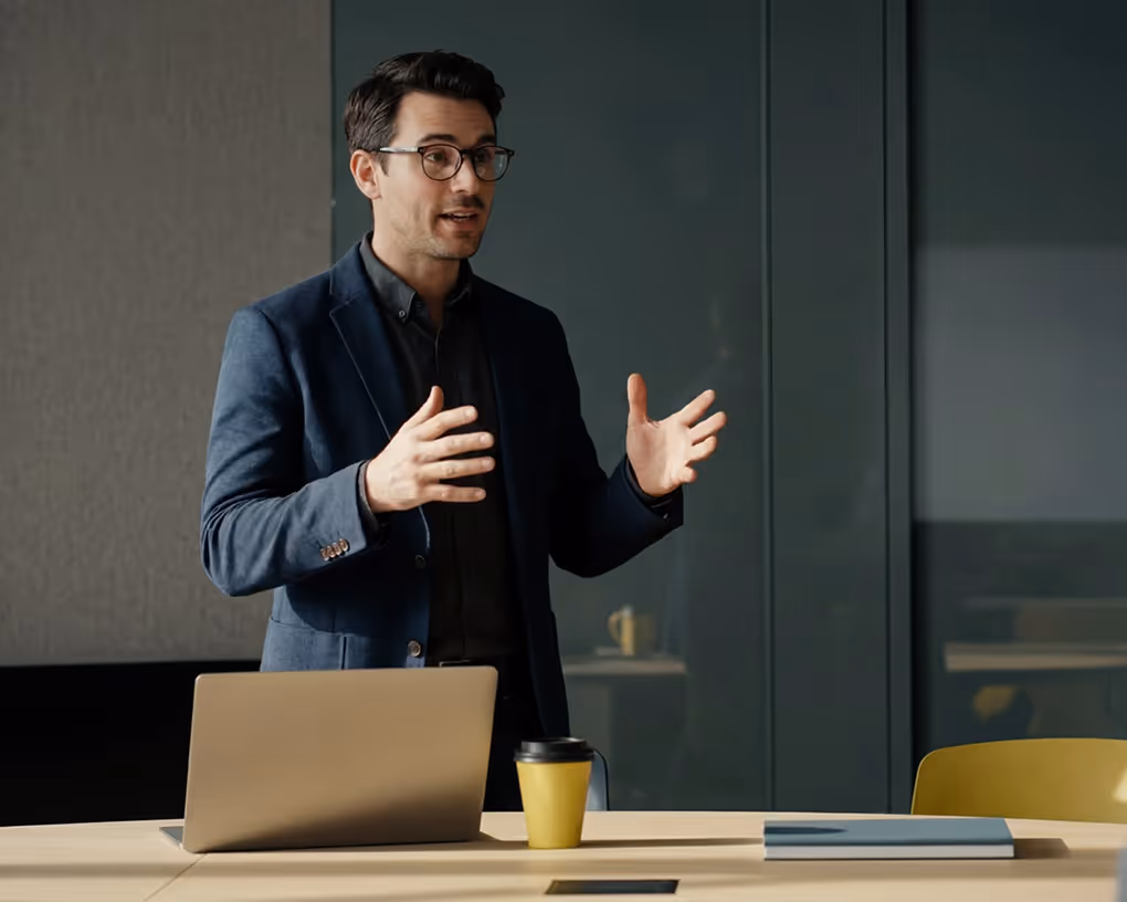 Man in glasses and blazer speaking and gesturing near laptop and yellow coffee cup on table.