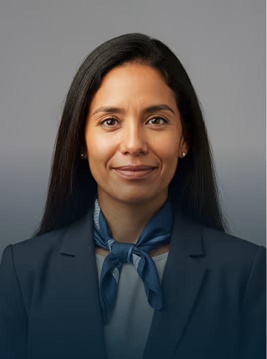 Smiling woman with long black hair wearing a dark blazer and blue neck scarf on gray background.