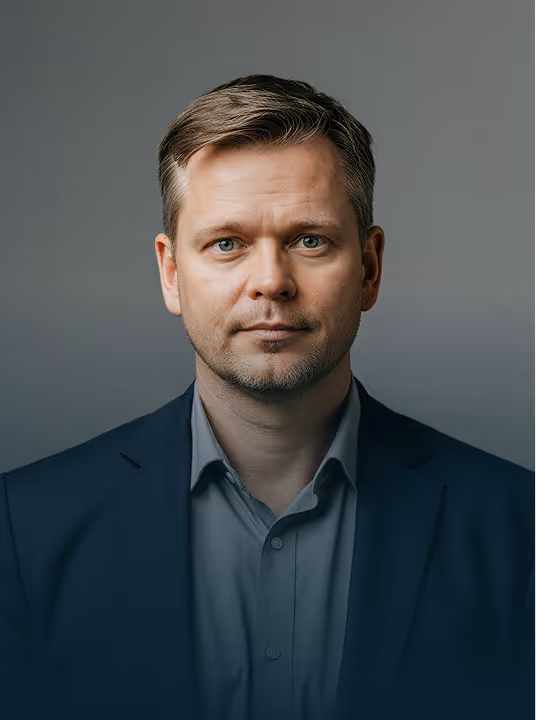 Portrait of a man with short light brown hair and blue eyes wearing a dark blazer and gray shirt.