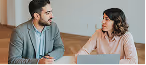Man and woman sitting at a table engaged in a serious conversation with a laptop in front of them.
